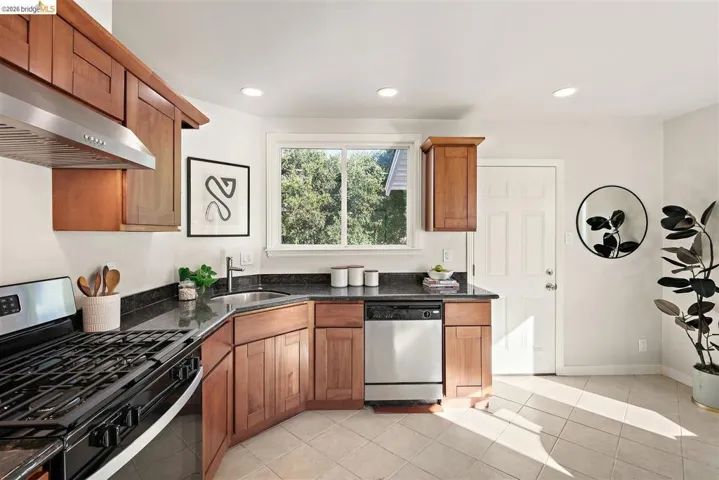Kitchen featuring wood cabinetry, dark countertops, a stainless steel range hood, stainless steel dishwasher, and tile flooring