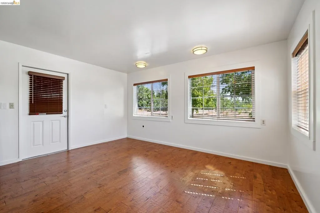 Spacious room featuring wood-finish flooring, multiple windows with blinds, a white door with a window, and two flush-mount ceiling lights