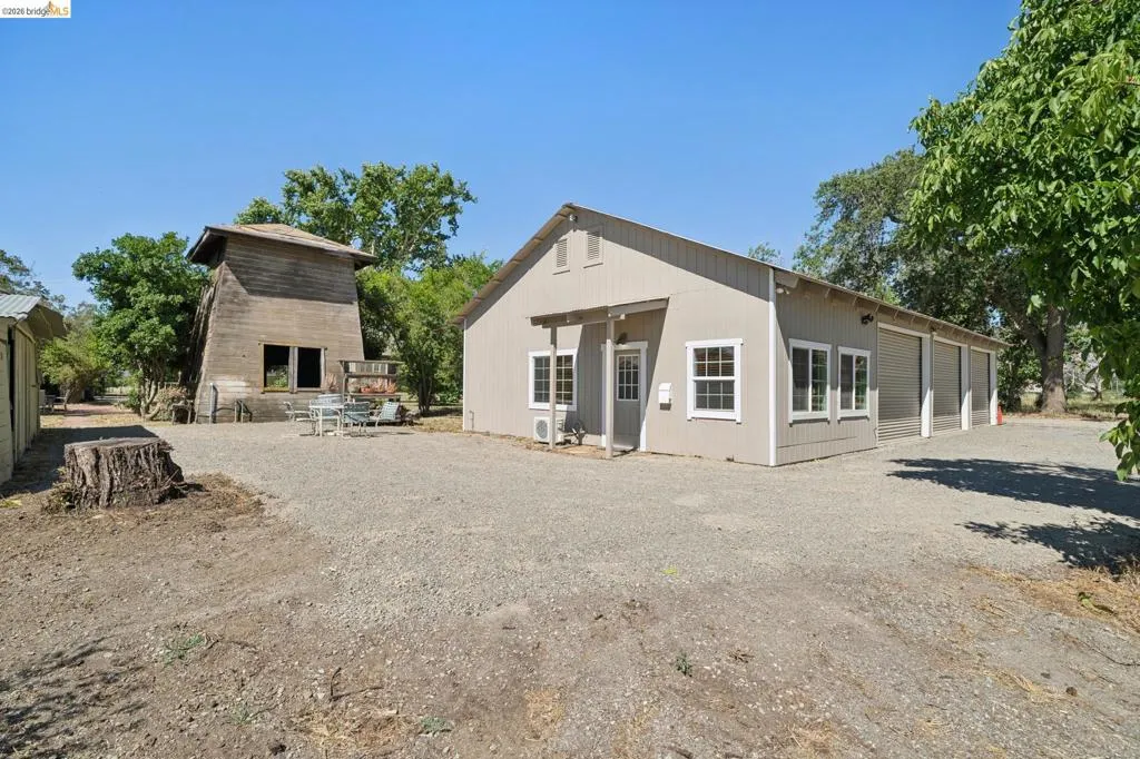 Expansive gravel area featuring a light-colored main building with multiple garage doors and windows, alongside a weathered wooden structure