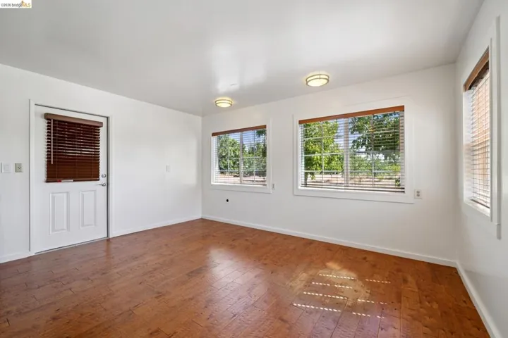 Spacious room featuring wood-finish flooring, multiple windows with blinds, a white door with a window, and two flush-mount ceiling lights