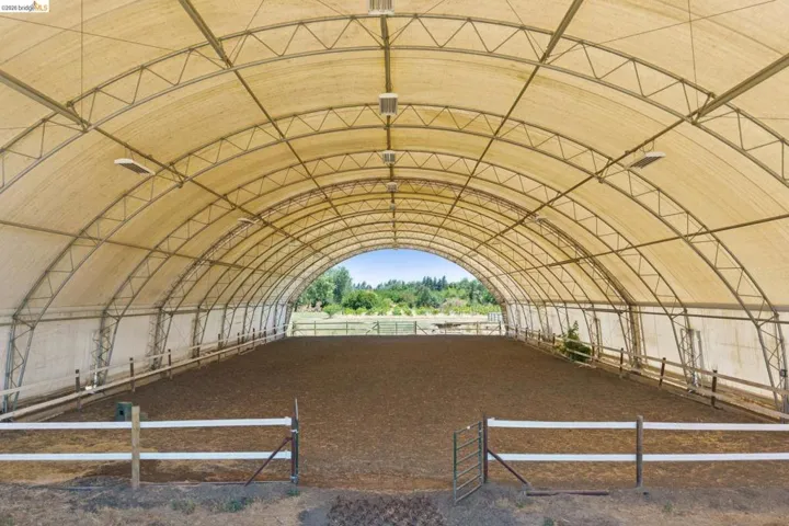 Expansive covered arena featuring a durable fabric roof with metal frame construction, recessed lighting, a perimeter fence with kickboard, and a soft ground surface