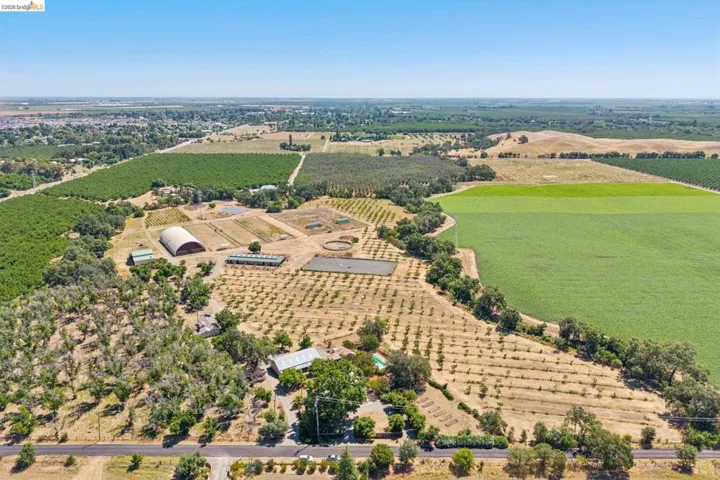Expansive agricultural property featuring cultivated fields, a large arched structure, multiple outbuildings, and mature trees