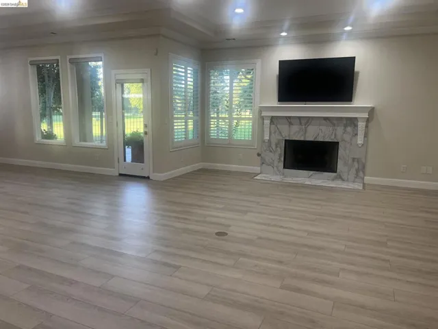 Spacious living area featuring light wood-finish flooring, a white mantel fireplace with a stone surround, and multiple windows with plantation shutters