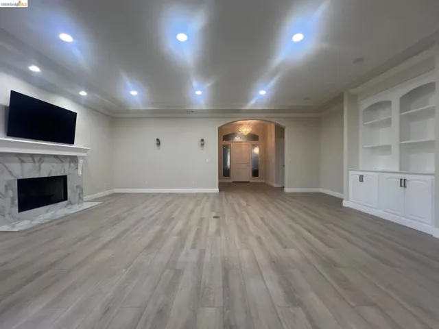 Spacious living area featuring wood-finish flooring, a white mantel fireplace with a stone-look surround, recessed lighting, and built-in white cabinetry with display shelving