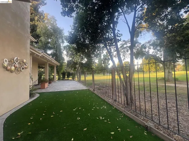 Backyard featuring artificial turf, expansive stone patio, covered porch, and black iron fencing bordering a golf course
