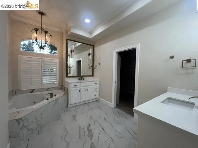 Spacious bathroom featuring a corner soaking tub with marble-look surround, plantation shutters, and a decorative chandelier
