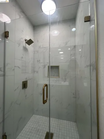 Glass-enclosed shower featuring large format white tile walls with subtle veining, a square rain showerhead, brushed gold fixtures, a recessed shower niche, and a tiled shower floor