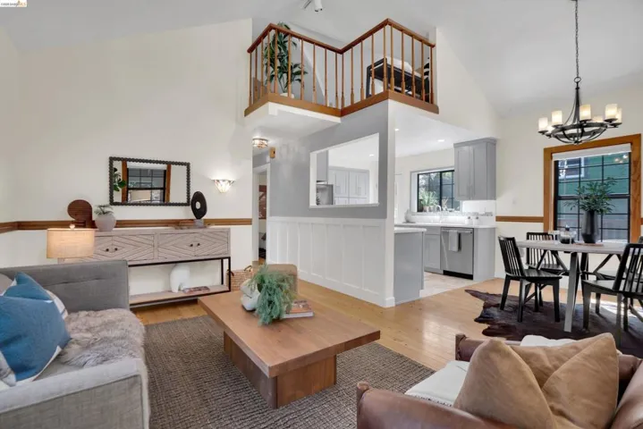 Vaulted ceiling living area featuring light wood-finish flooring, a built-in kitchen pass-through window, and an upper-level loft with wood railings
