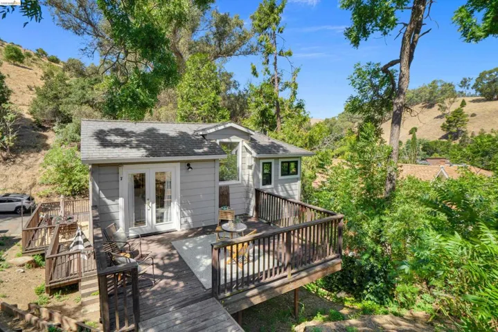 Elevated wood-plank deck with railing, featuring a light grey exterior with white trim, double glass-paned doors, and surrounding mature trees