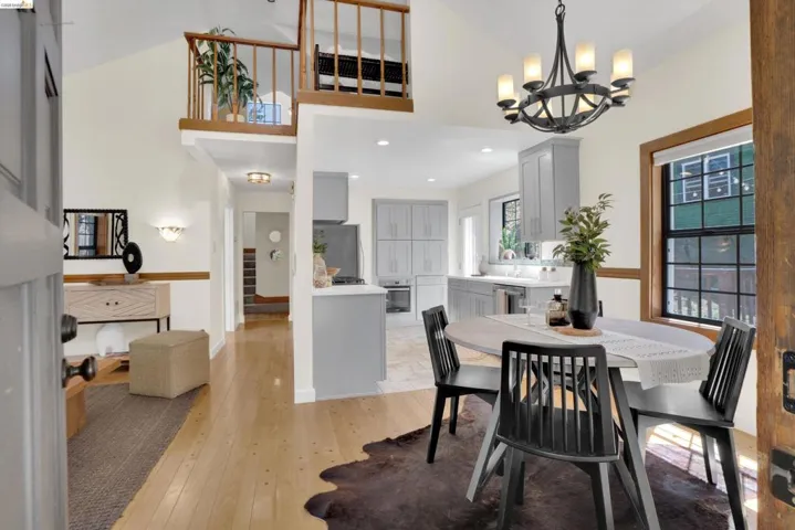Open-concept living space featuring wood-finish flooring, a contemporary kitchen with light grey cabinetry, a dining area with an overhead chandelier, a lofted area with wood railings, and a hallway leading to another section of the property