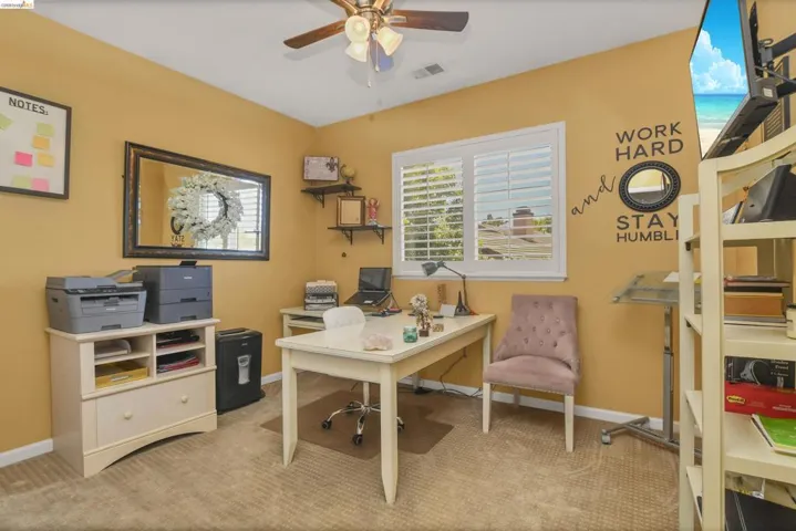 Carpeted room featuring a ceiling fan with integrated lighting, a rectangular window with white plantation shutters, built-in wall shelving, and a built-in white desk