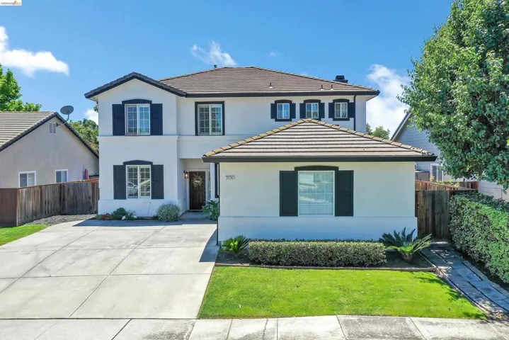 Contemporary stucco exterior featuring dark shutters, a concrete driveway, a tiled roof, a manicured lawn, and mature landscaping