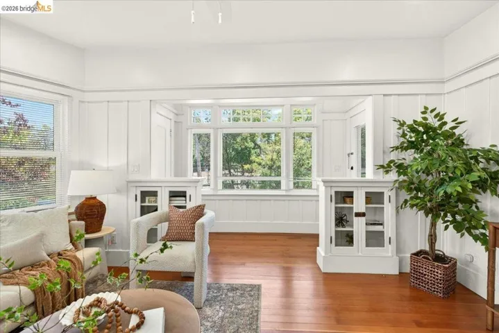 Hardwood flooring throughout featuring white board and batten wall paneling, built-in display cabinetry with glass doors, a large bay window, and additional window with horizontal blinds