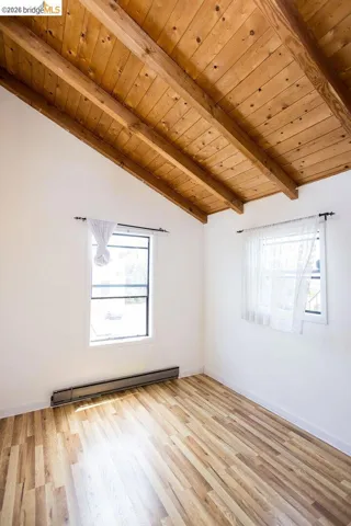 Vaulted ceiling with exposed wood beams, bright white walls, two windows, wood-finish flooring, and baseboard heating