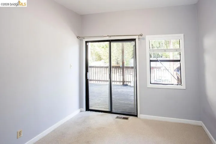 Spacious room featuring light gray walls, plush carpeting, a large sliding glass door opening to a private wooden deck, and an additional window providing natural illumination