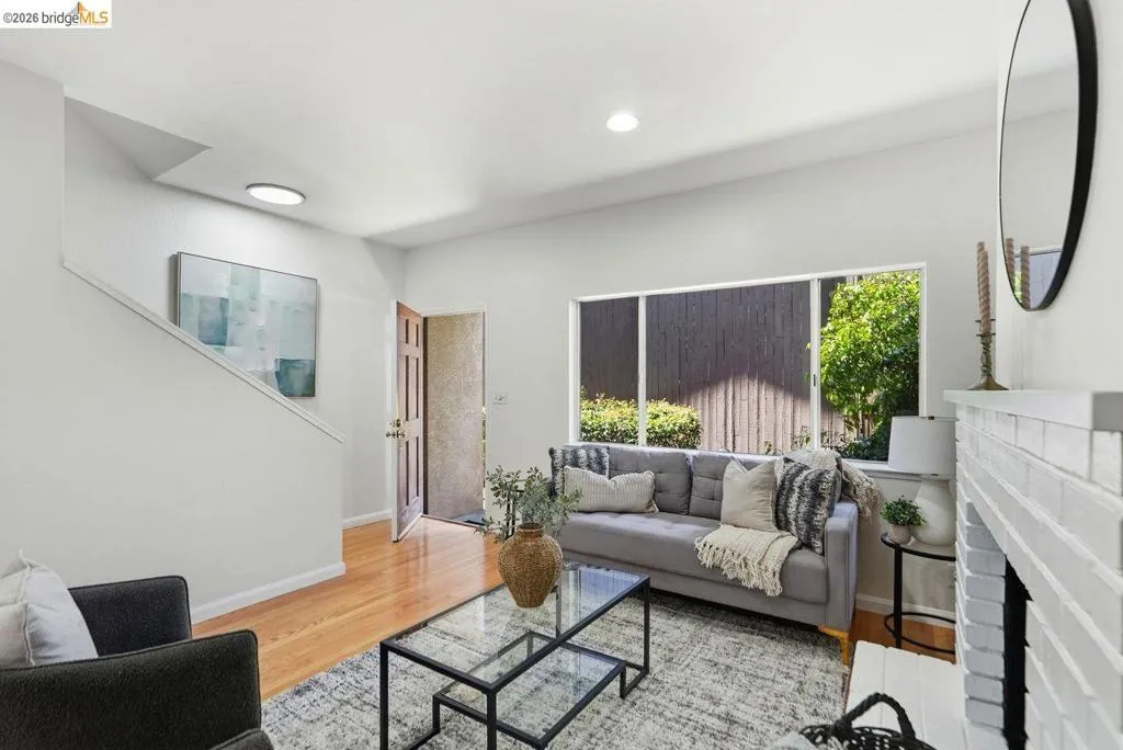 Bright living space featuring natural wood flooring, a white brick fireplace, and a large window overlooking exterior greenery