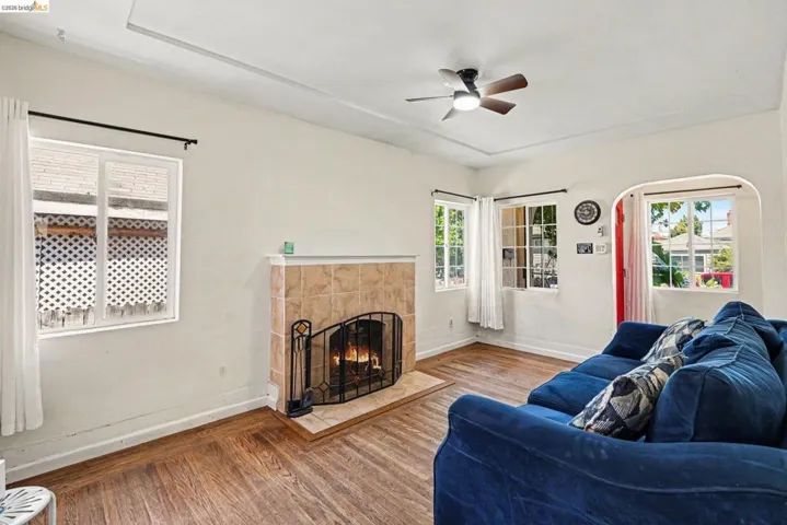 Living area featuring wood-finish flooring, a tiled fireplace with a hearth, multiple windows for natural light, a ceiling fan, and a tray ceiling
