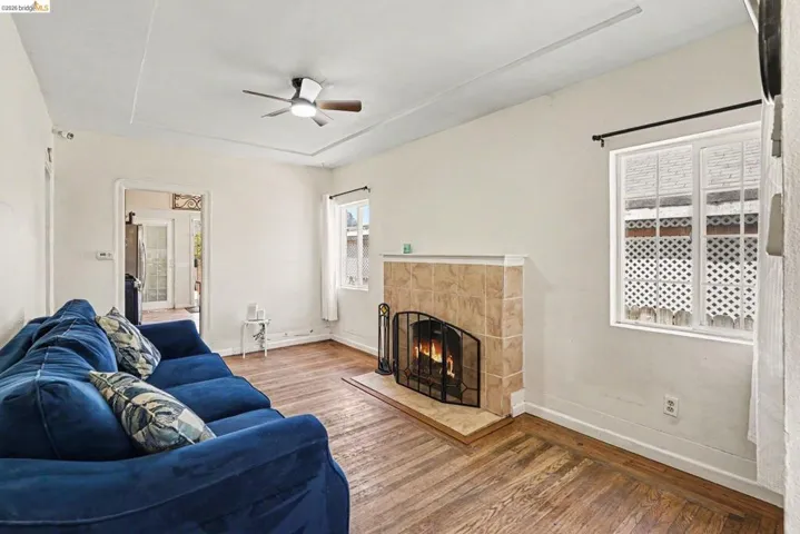 Living area featuring wood-finish flooring, a tiled fireplace with a metal screen, two windows with white trim, and a ceiling fan with integrated lighting