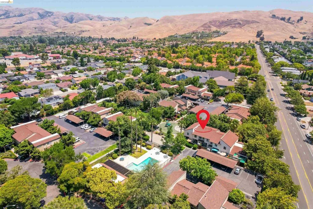 Aerial perspective showcasing a multi-unit property with a red pin marker, featuring a swimming pool, covered parking areas, mature landscaping, and proximity to a tree-lined main thoroughfare