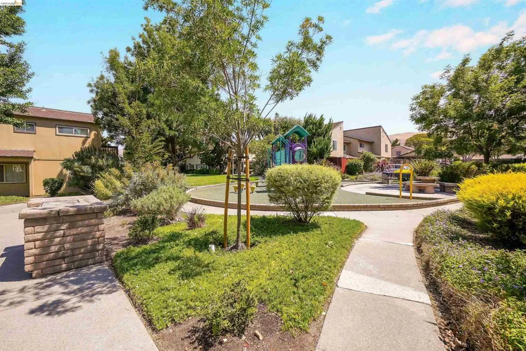 Community green space featuring a playground with colorful equipment, a circular turf area, and surrounding mature landscaping