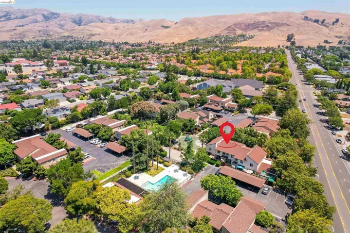 Aerial perspective showcasing a multi-unit property with a red pin marker, featuring a swimming pool, covered parking areas, mature landscaping, and proximity to a tree-lined main thoroughfare