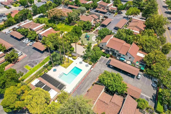 Community-style property featuring multiple buildings with red tile roofs, a central swimming pool with surrounding patio, a playground area, and covered parking structures