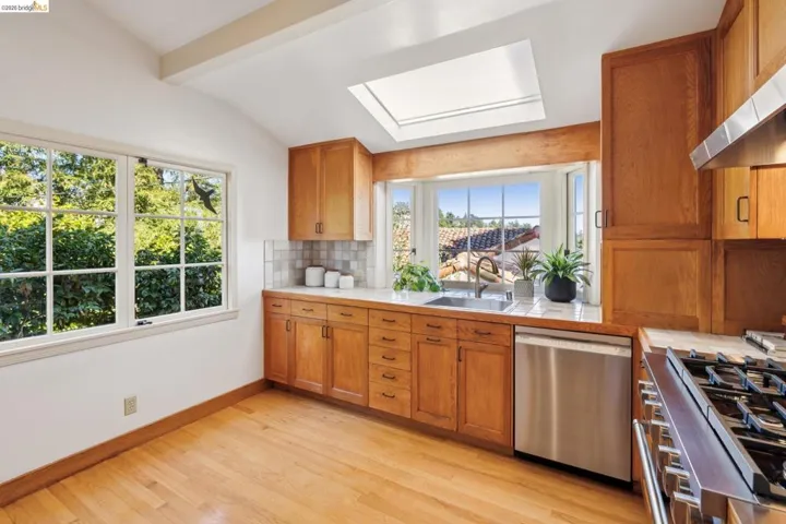 Kitchen featuring wood-finish flooring, wood cabinetry, a skylight, a large multi-pane window, and a stainless steel dishwasher