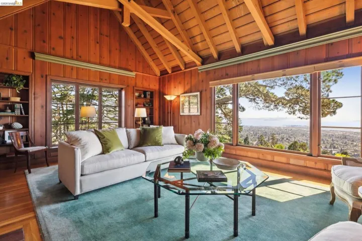 Spacious living area featuring extensive wood paneling, an exposed beam vaulted ceiling, large picture windows with Bay views, built-in shelving, fireplace and hardwood floors