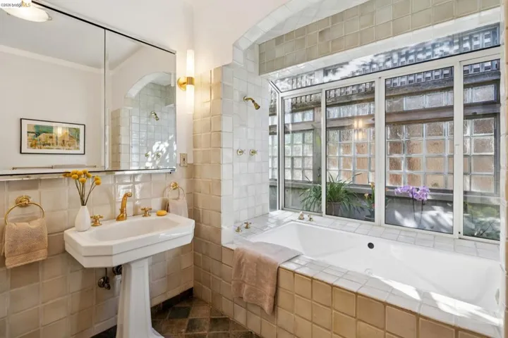Tiled bathroom featuring a built-in soaking tub, large window with glass block detailing, pedestal sink, and gold-finish fixtures