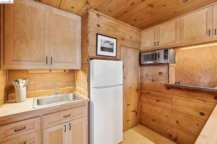 Compact kitchen featuring wood-paneled walls and ceiling, light wood cabinetry, a stainless steel sink, and a white refrigerator