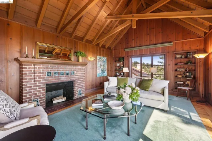 Spacious living area featuring extensive wood paneling, an exposed beam vaulted ceiling, large picture windows with Bay views, built-in shelving, fireplace and hardwood floors