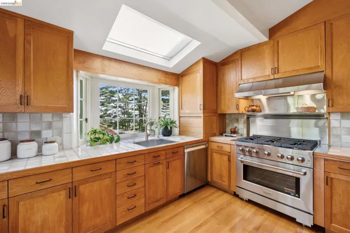 Kitchen featuring natural wood cabinetry, light tile countertops, a stainless steel range with matching hood, a single basin sink, and wood-finish flooring