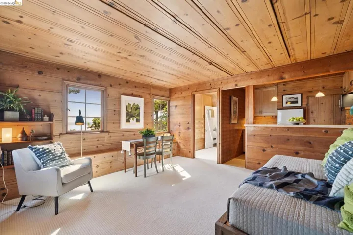 Knotty pine paneling throughout, light-toned carpet, a built-in shelf unit, multiple windows providing natural light, and a compact kitchenette with pendant lighting