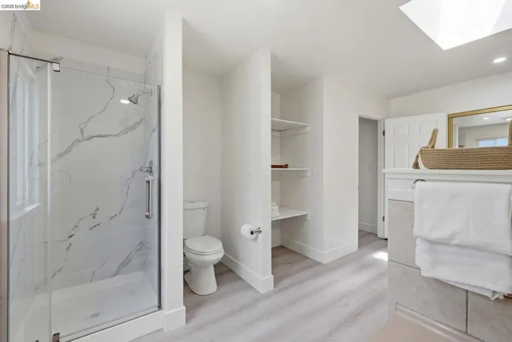 Modern master bathroom featuring a glass-enclosed shower with a marble-patterned surround, light wood-finish flooring, built-in shelving, a skylight, and a vanity with a mirror
