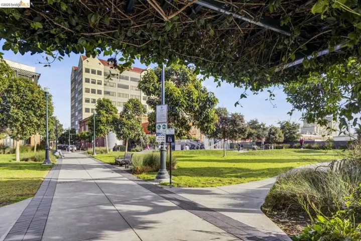 Landscaped park area featuring a concrete pathway with brick accents, mature trees, and open green spaces