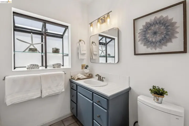 Bathroom featuring a casement window with glass shelving, a vanity with a white countertop and integrated sink, a rectangular mirror, a three-bulb light fixture, and a toilet