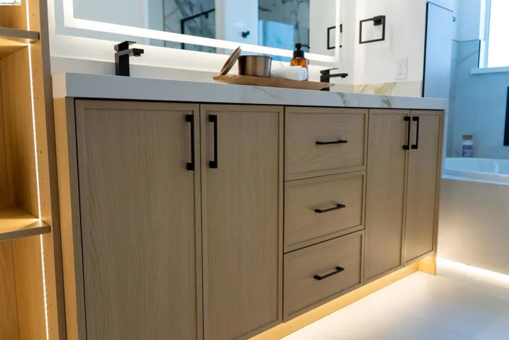 Double vanity with light wood-finish cabinetry, black hardware, and under-cabinet lighting