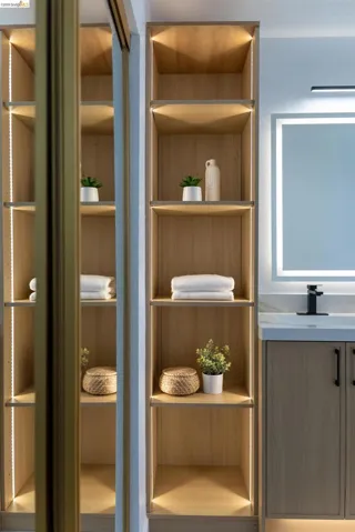 Built-in shelving with integrated lighting, wood-finish cabinetry, a modern vanity with a rectangular illuminated mirror, and a matte black faucet