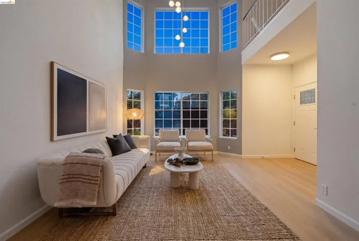 Double-height living space featuring large grid-pattern windows, light wood-finish flooring, and an upper-level balcony with white railing
