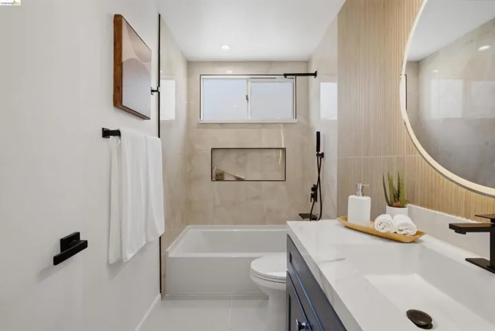 Modern bathroom featuring a single vanity with a white countertop and dark blue cabinetry, a back-lit round mirror, a white bathtub, and light-toned tiled walls with a built-in niche