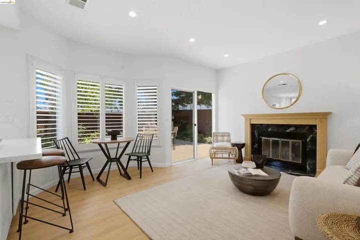 Open-concept living area featuring light wood-finish flooring, a traditional fireplace with a dark stone surround and wood mantel, white plantation shutters, and recessed lighting