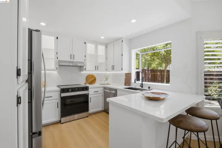 Bright kitchen featuring white cabinetry with black hardware, stainless steel appliances, light-toned wood-finish flooring, and a large window overlooking lush greenery