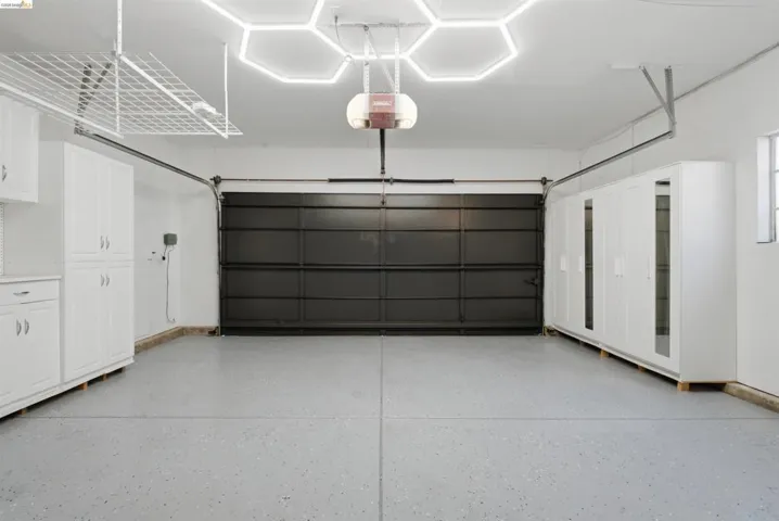 Spacious two-car garage featuring extensive built-in white cabinetry, a durable epoxy floor, overhead storage racks, modern hexagon-patterned ceiling lighting, and a dark-paneled garage door