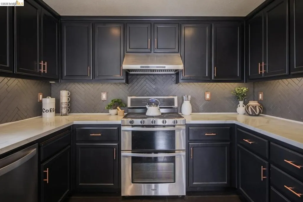 U-shaped kitchen featuring dark cabinetry with copper-toned hardware, light-toned countertops, and a herringbone-patterned backsplash