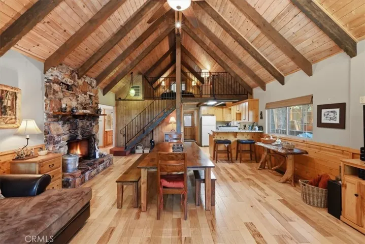 Dining area with vaulted wood ceilings, stone fireplace, staircase, and kitchen beyond.