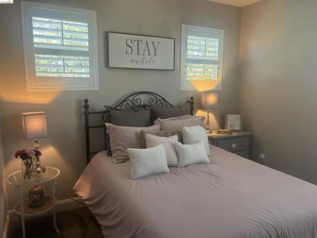 Bedroom featuring two windows with white plantation shutters, a dark-toned metal headboard, a gray painted nightstand with a single drawer and cabinet, and light gray wall paint