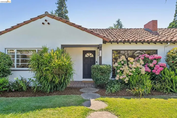 White stucco exterior featuring a terra cotta tile roof, dark-framed entry door, front picture window, and mature landscaping with hydrangeas