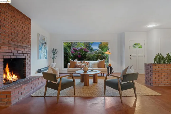 Living area featuring a brick fireplace, a large picture window with extensive outdoor greenery, and wood-finish flooring