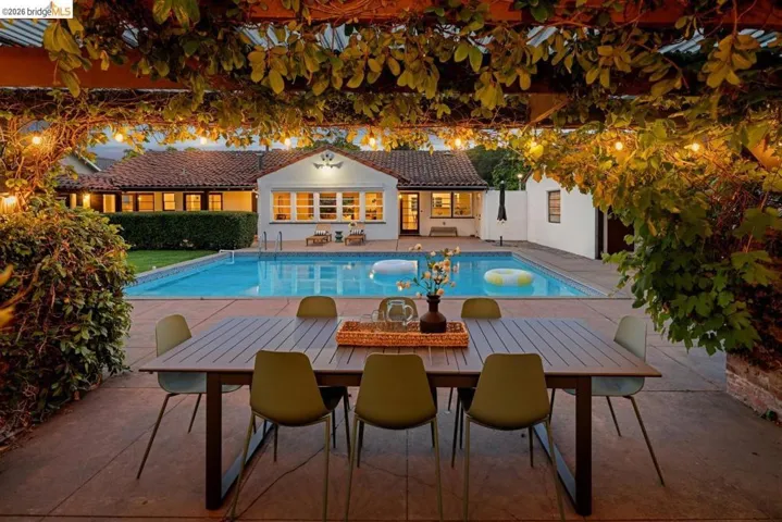 Patio dining area featuring a wood-finish table and seating, under a pergola with lush foliage and string lighting