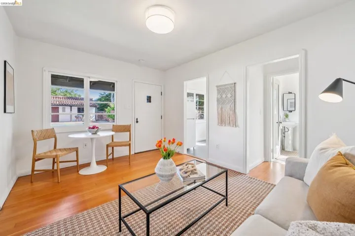Bright living area featuring wood-finish flooring, white walls, a large window, a ceiling-mounted light fixture, and an open doorway leading to a bathroom with a pedestal sink